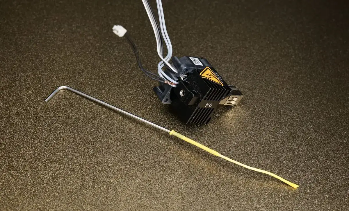 Resíduo de filamento removido do interior de um hotend de impressora 3D ao lado da ferramenta de limpeza